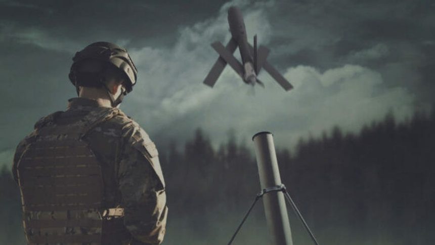 A soldier stands near a launch tube as a military drone flies overhead in a battlefield setting.