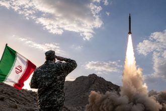 Iranian soldier saluting beside the Iran flag during a missile launch.