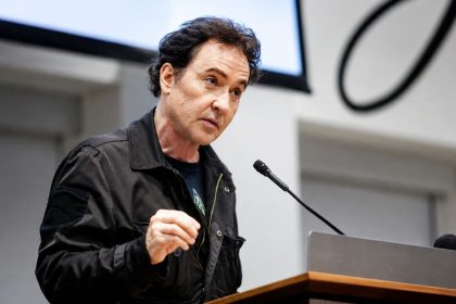 John Cusack delivers a speech at a podium during a public event. Image Credit: Bloomberg/Getty Images.