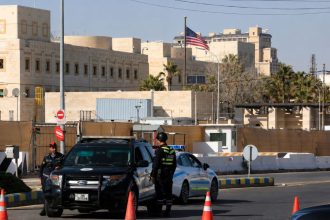 Police officers secure the area outside the U.S. Embassy in Amman, Jordan, with embassy buildings and the U.S. flag visible in the background.
