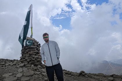 Irish-British trail runner Alexander Mark Harris poses beside a Pakistani flag at a mountain summit.