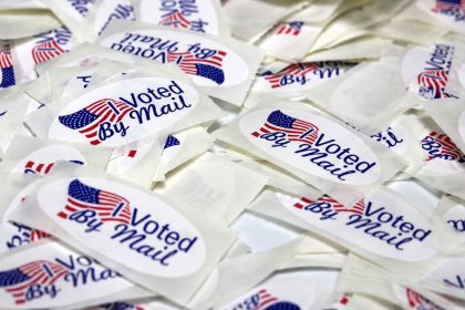 “Voted By Mail” stickers are pictured in a pile, featuring American flag designs.