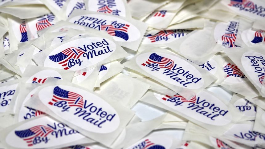 “Voted By Mail” stickers are pictured in a pile, featuring American flag designs.