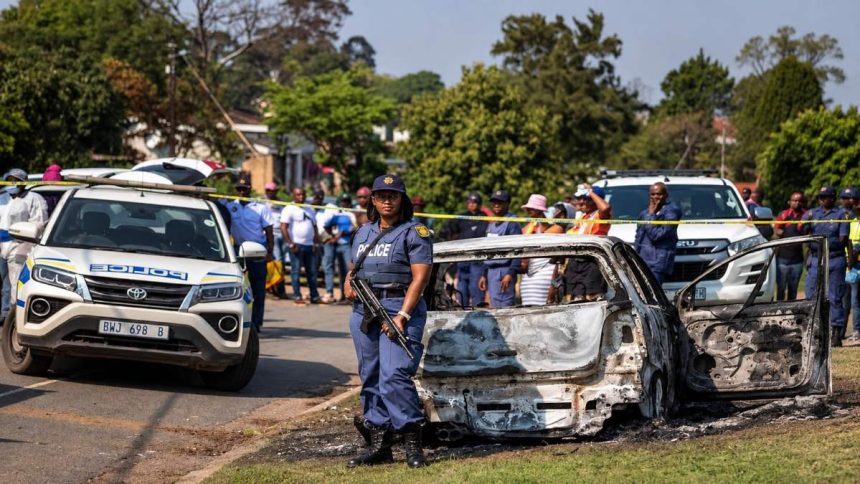South African police officer standing beside a burned-out car at a crime scene.