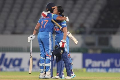 India women's cricket players in blue jerseys embrace on the field while holding their bats and helmets.