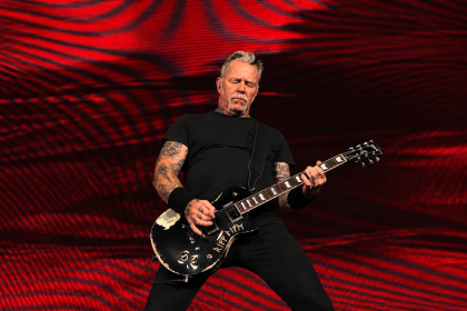 Metallica frontman James Hetfield playing a black electric guitar during a live performance, wearing a black T-shirt and black trousers.