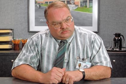Richard Riehle wearing glasses, a striped shirt and tie while seated at an office desk.