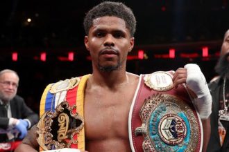 Shakur Stevenson wearing multiple championship belts inside the boxing ring.