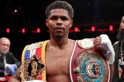 Shakur Stevenson wearing multiple championship belts inside the boxing ring.