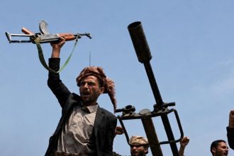 Armed Houthi fighters raise a rifle and fists beside a mounted weapon in Yemen.