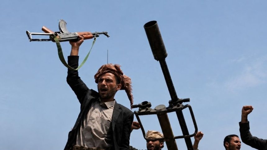 Armed Houthi fighters raise a rifle and fists beside a mounted weapon in Yemen.